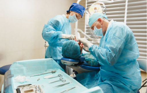 A dentist and dental assistant wearing masks perform a dental procedure on a patient with their equipment laid out in from of them