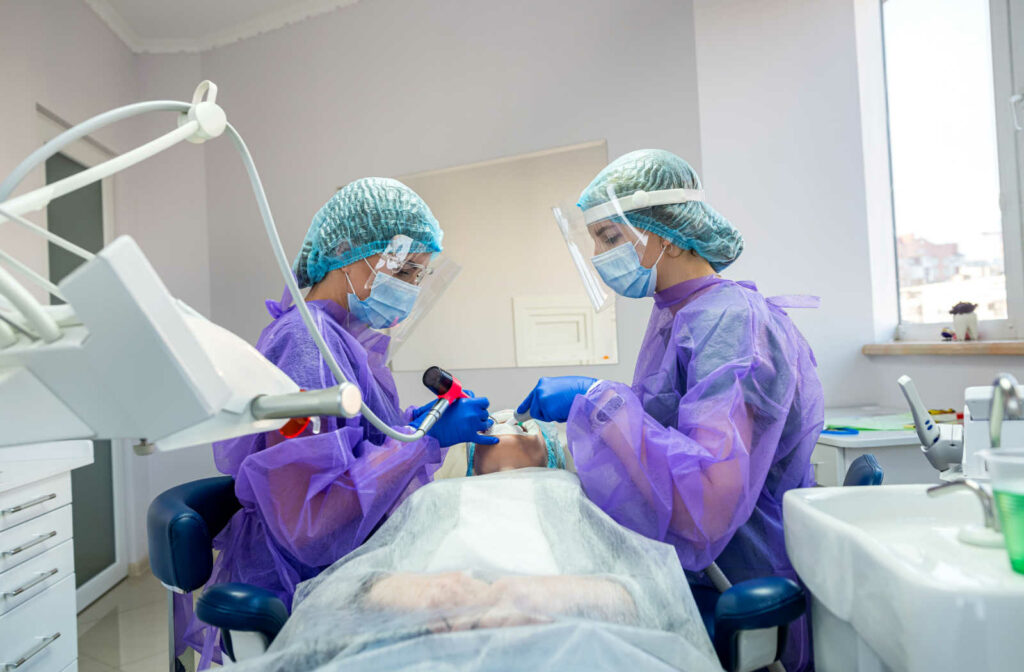 A dental team working together in a well-lit dental office. The dentist is examining a patient's teeth while the dental assistant stands by, holding a tray of dental tools. The patient is reclined in a dental chair wearing protective eyewear. The room is equipped with dental equipment such as a light, a dental unit, and a computer monitor.