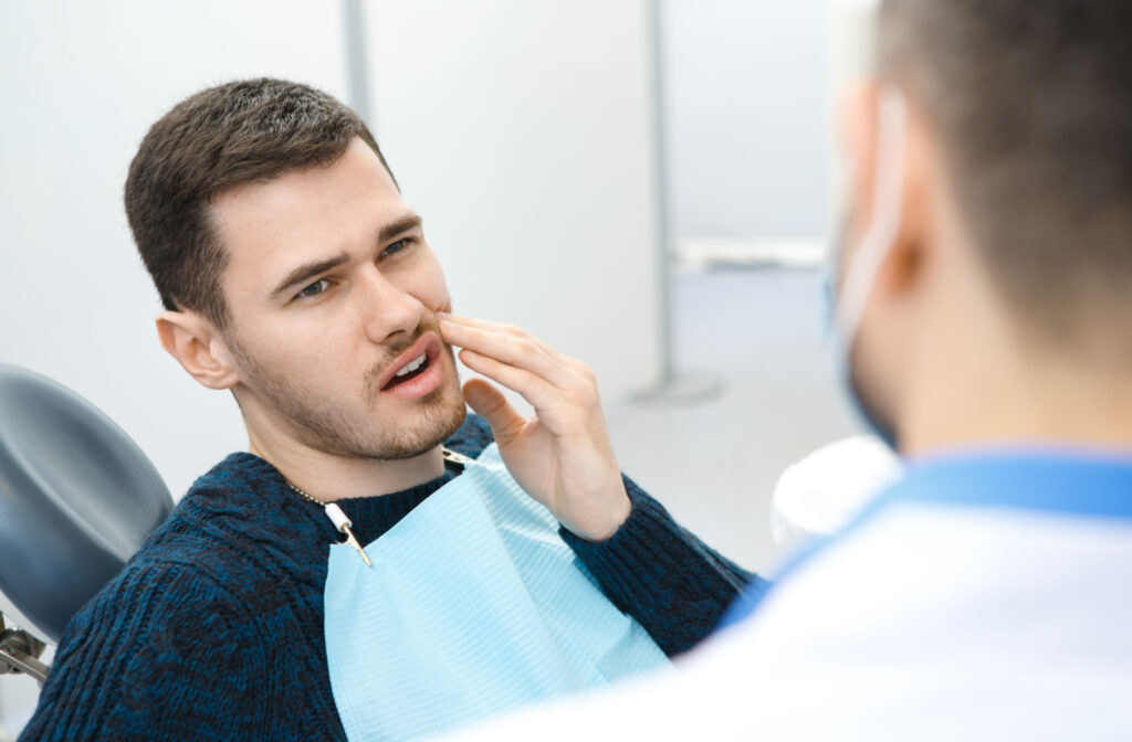 A young man with dental bib sitting in a dentist office holding his left check with his left hand while talking to his dentist.
