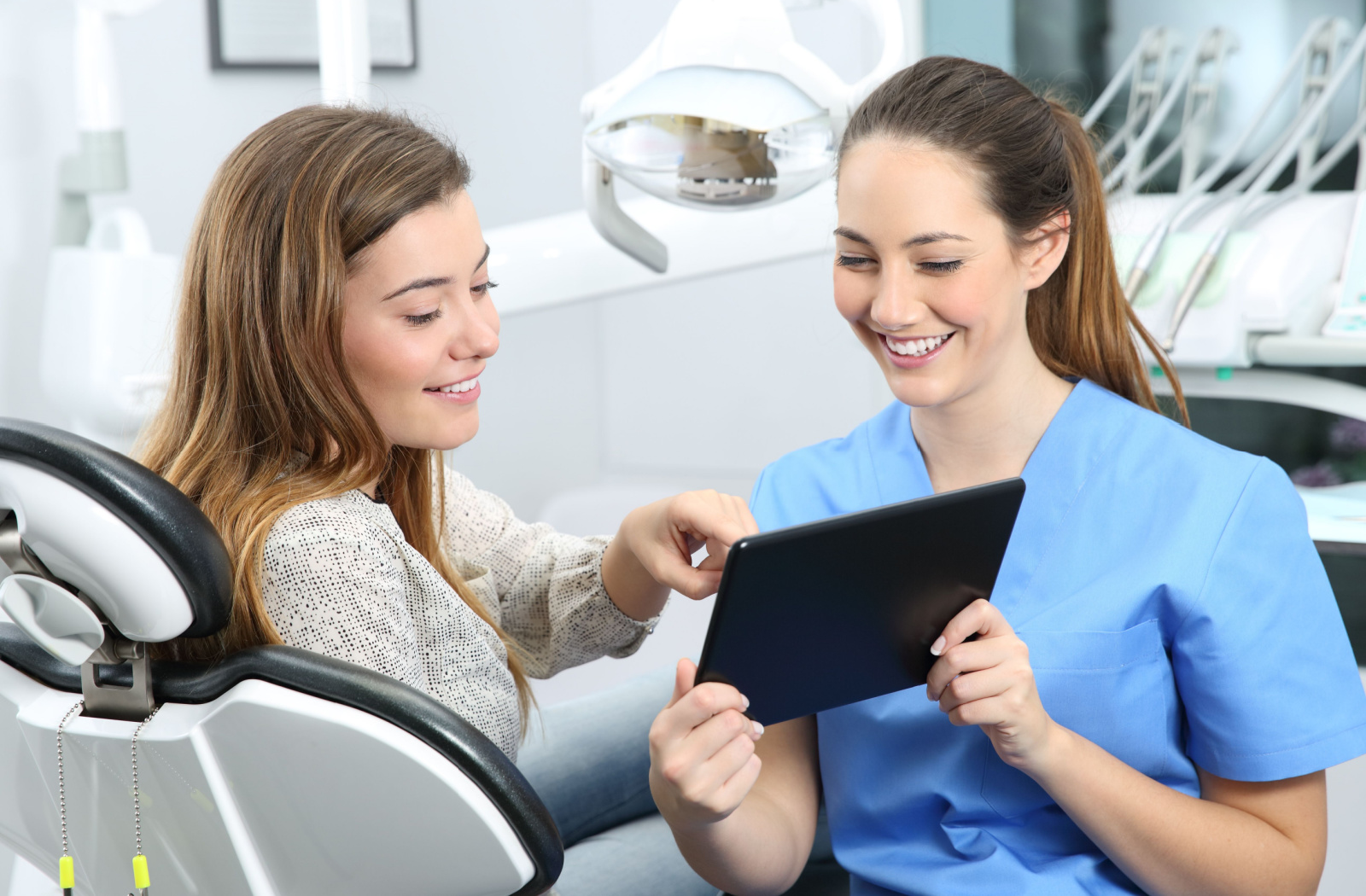 A female dentist in blue scrubs holding a clipboard and smiling at her patient in the dental chair.