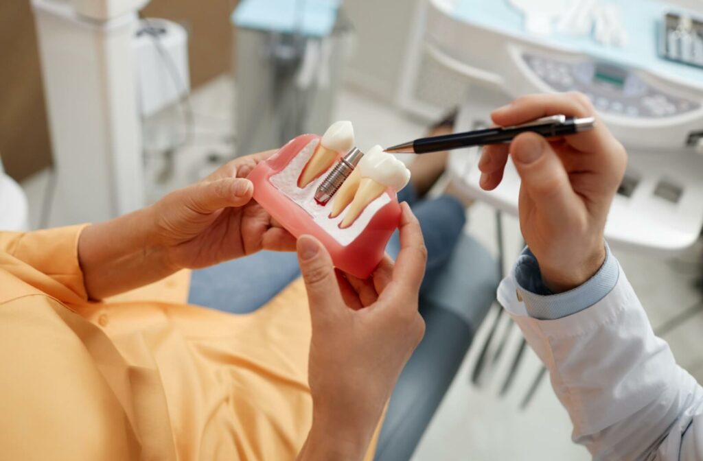 Close-up of a patient's hands holding an enlarged model of teeth in gums with 1 dental implant. A dentist points to one tooth with a pen.