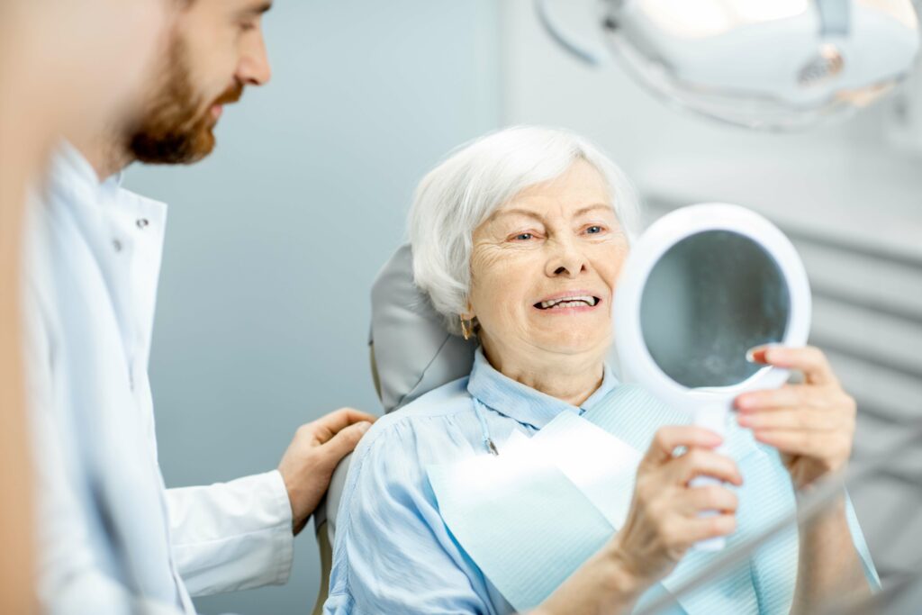 An older woman sitting in a dental chair and admiring her smile in a handheld mirror as her dentist looks on.