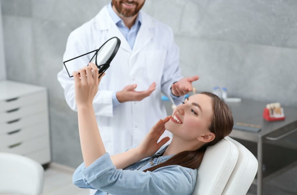 A woman sitting in a dental chair smiles while holding a mirror to examine her teeth during a dental appointment.