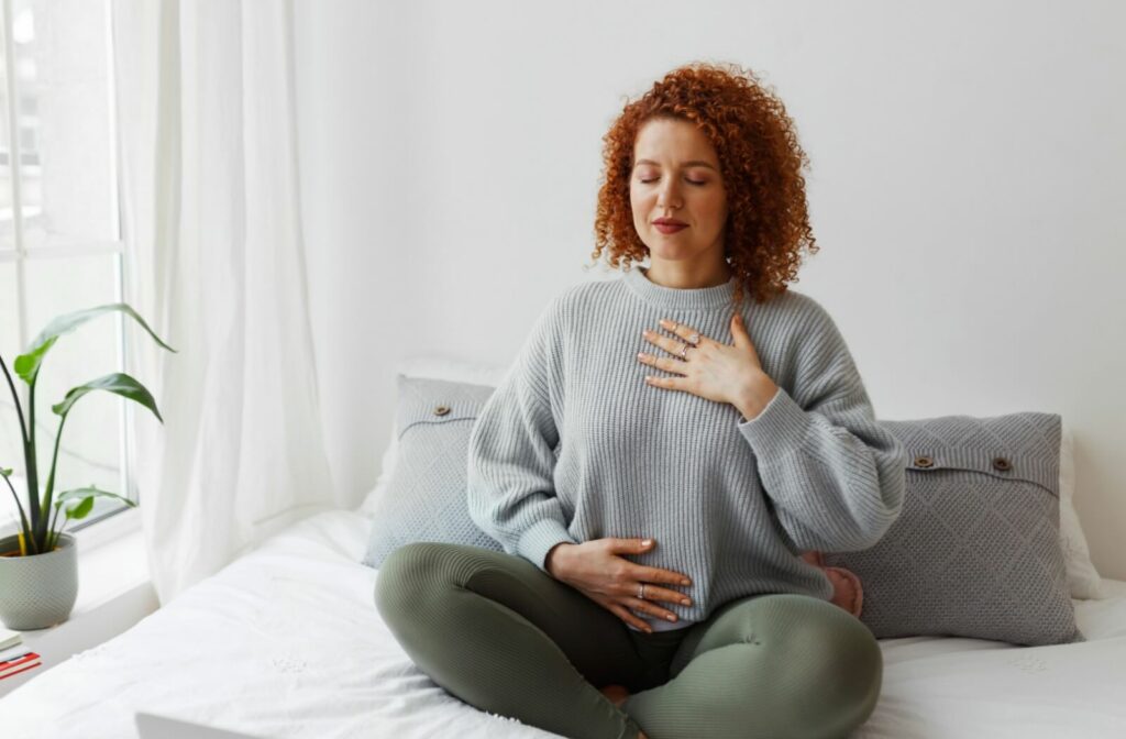 A person in a sweater and yoga pants sits cross-legged on a bed and practices deep breathing relaxation techniques.