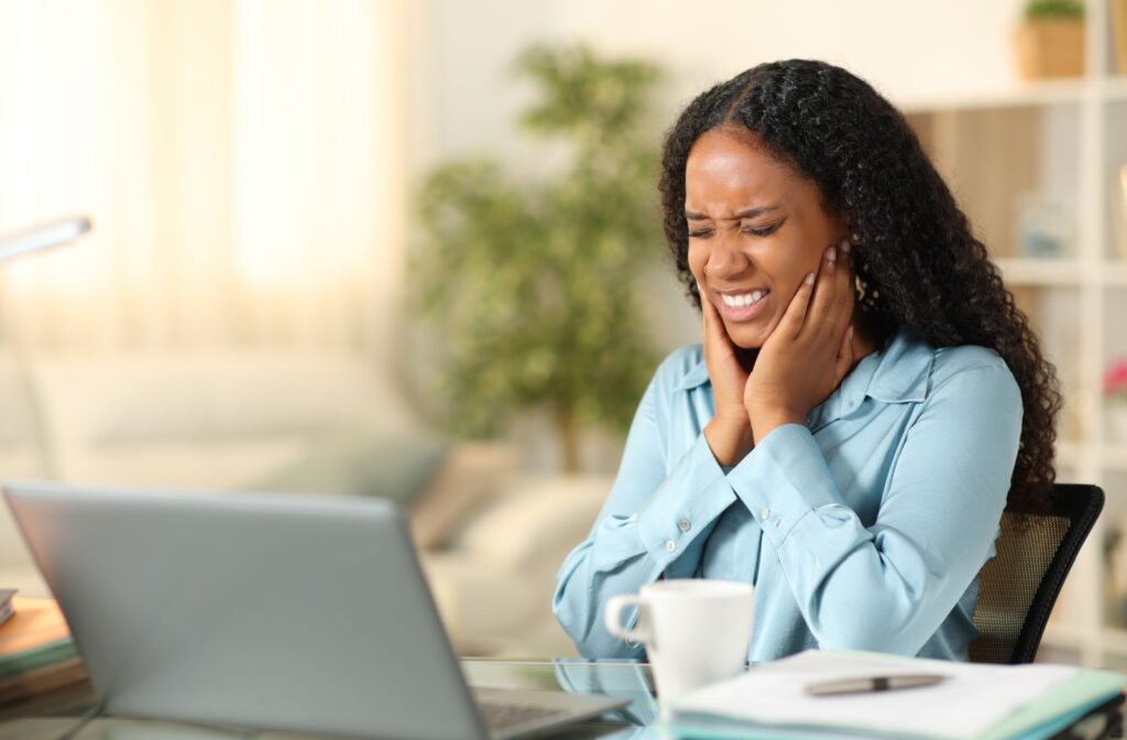 A woman sits at a table in front of a laptop in a brightly lit room, her hands clutching either side of their jaw due to the discomfort of a TMJ-related toothache.