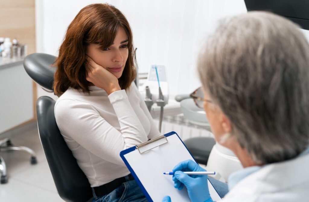 A patient sitting in an exam chair, holding their jaw, speaks with an oral surgeon about tooth pain while the surgeon discusses TMJ-related symptoms.