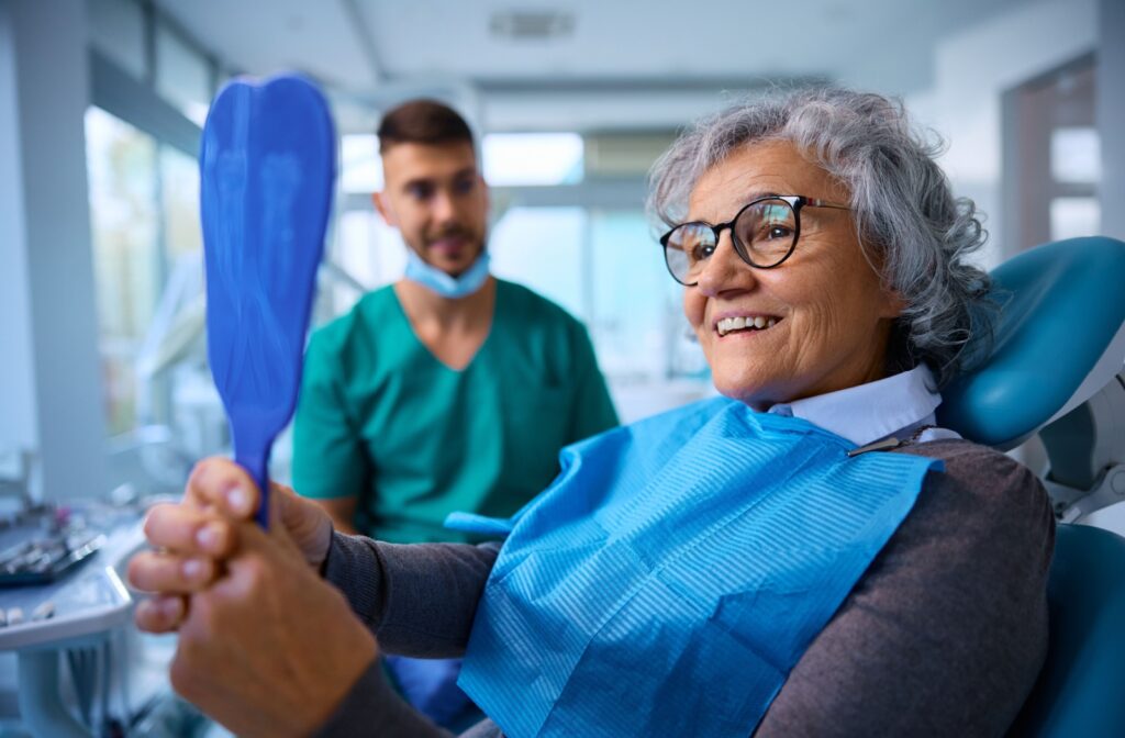 A patient smiles as they examine their new dental implants in a handheld mirror, sitting comfortably in a dental chair. In the background, their oral surgeon looks on while smiling.